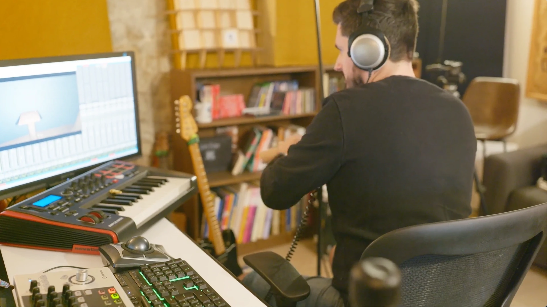 Sound Designer Romain Raynal, wearing a studio headset, is working on a video game project at his audio production station. The main screen displays audio software (DAW) and a gaming scene. On his desk, we can see a MIDI Novation Impulse keyboard, a Cubase CC121 control interface and a trackball.