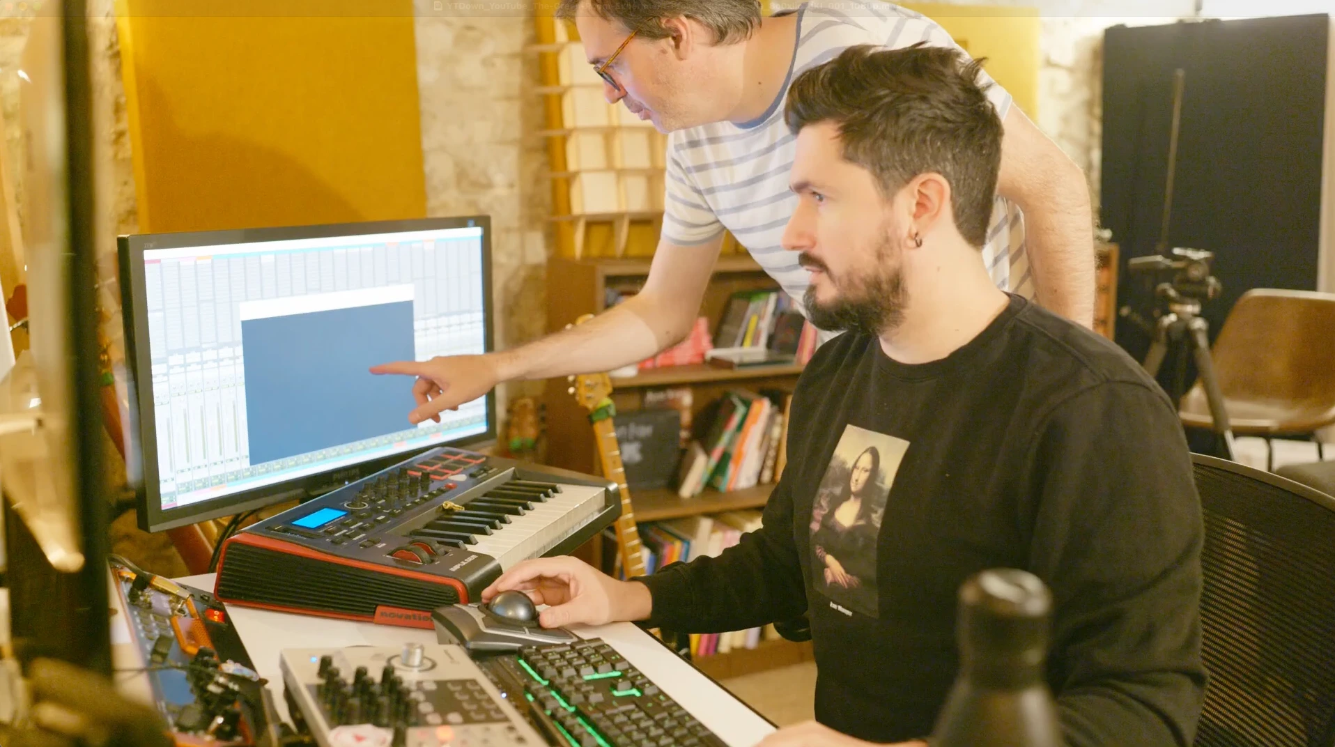 The Sound Designer Romain Raynal sitting at the office, collaborates with a colleague who points to a specific audio plugin (presumably FabFilter Pro-Q) on the screen displaying a DAW. The studio’s full equipment is visible, including a MIDI keyboard and audio interface.